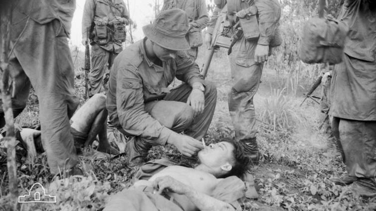An Australian soldier gives a wounded North Vietnamese Army soldier a cigarette while waiting for an evacuation helicopter. Photographer: Christopher John Bellis. Image courtesy of the Australian War Memorial BEL/69/0394/VN.