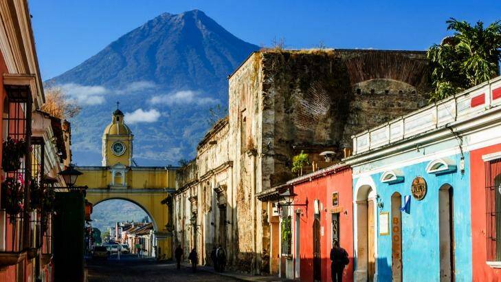 Locals walk to work along Antigua's famous cobblestoned street lined with painted shops, restaurants & hotels. Agua volcano looms behind Santa Catalina Arch, a landmark in this Spanish colonial town & UNESCO World Heritage Site. Photo: iStock